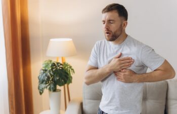 A young man is standing at home and holding his chest. Feels severe pain, shortness of breath, panic attack, heart attack, stroke.